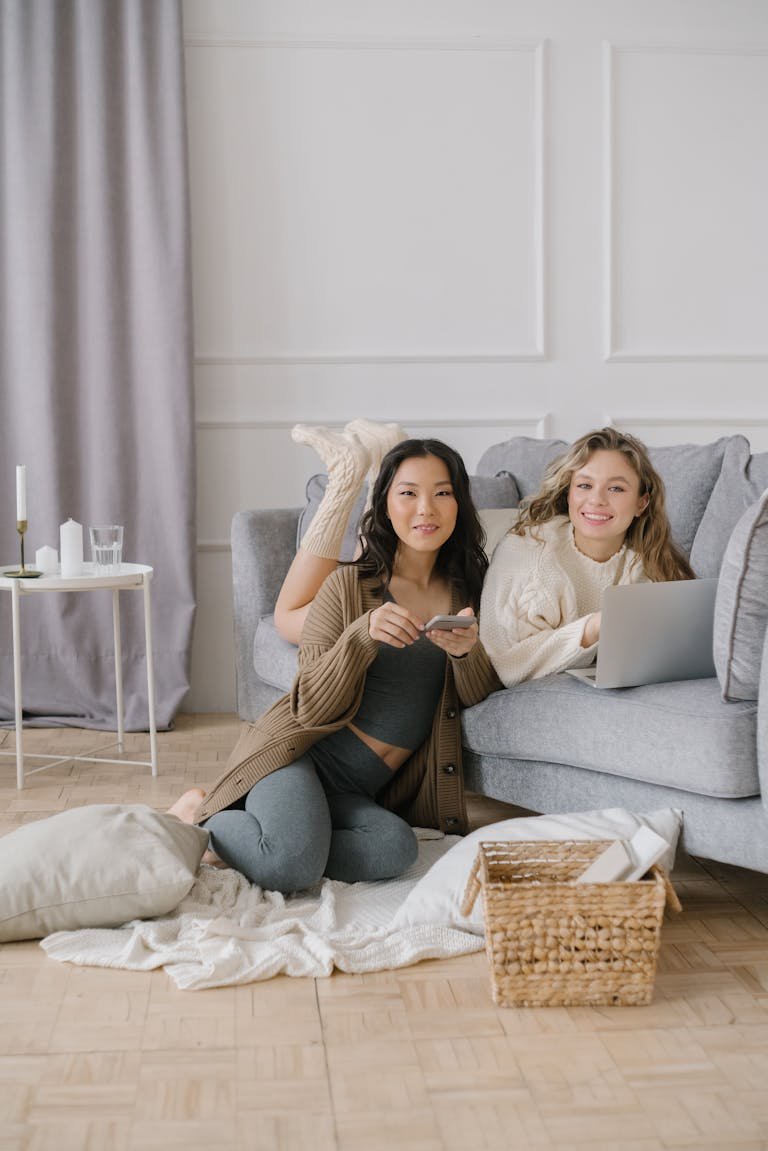 Two friends relaxing at home using a laptop and smartphone, showcasing comfort and connectivity indoors.