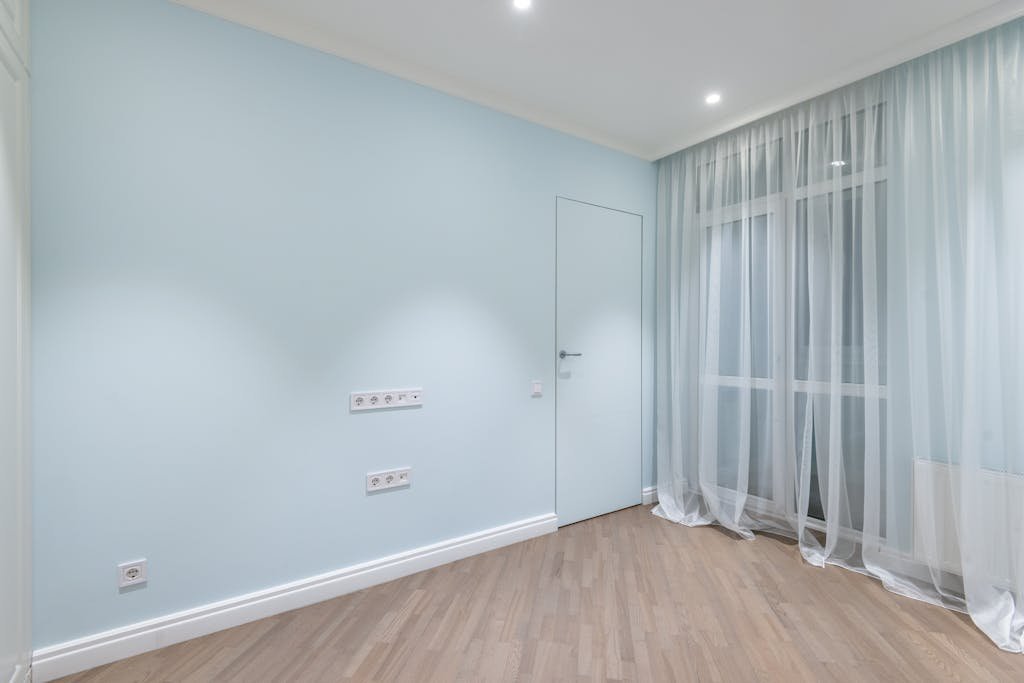 A modern, minimalist empty room with light blue walls and sheer curtains, offering a clean and spacious atmosphere.