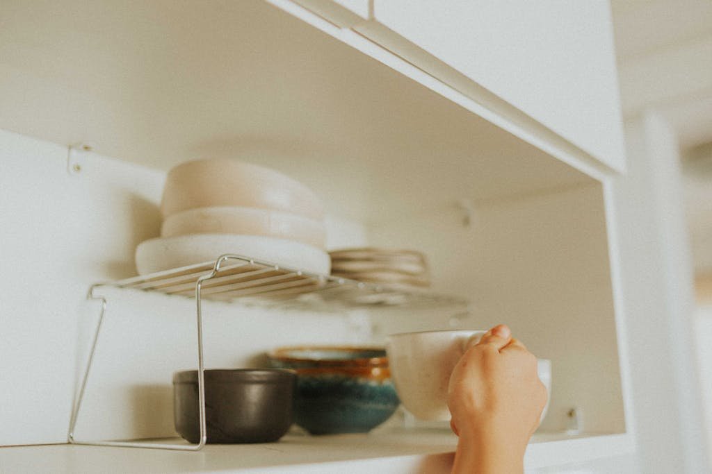 A person reaches into a kitchen cabinet to grab a mug, surrounded by neatly stacked dishes.