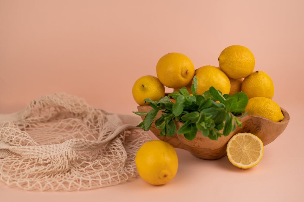 Aesthetic composition of fresh lemons and mint leaves in a wooden bowl with a mesh bag on a peach backdrop.