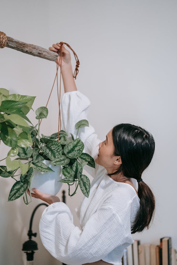 An adult Asian woman with long black hair adjusts a hanging potted plant indoors, showcasing a casual white outfit.