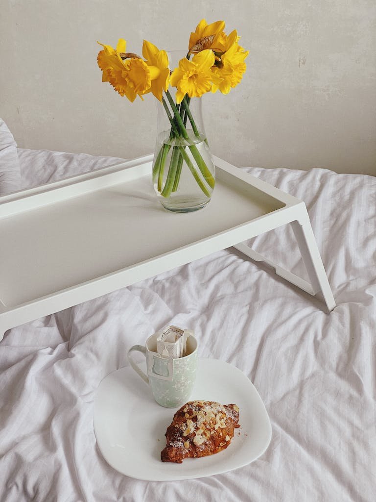 From above of cup of drip coffee with paper filter and plate with fresh croissant served on soft bed near tray with vase of yellow Narcissus jonquilla flowers