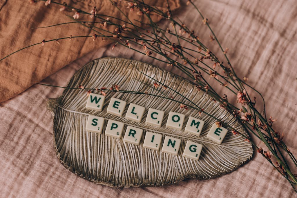 Letter tiles spelling 'Welcome Spring' on a decorative leaf with delicate flowers, evoking a spring theme.